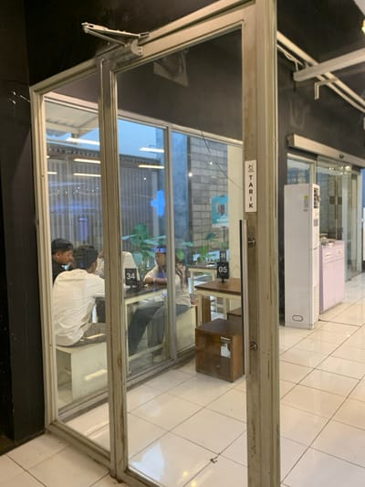 The image shows a view through a glass door, looking into a cafe in Malang, Indonesia, located at latitude -7.941 and longitude 112.608. There are four people seated at two tables, engaged in conversation. One man is wearing a white shirt, another man is wearing a white shirt, and a woman is wearing a white shirt. The tabletops are marked with numbers 34 and 35, indicating seating arrangements. There are also visible signs and other furniture. The cafe's floor is tiled in white. The environment is bright and well-lit. The door is partially open, with a sign that says "TARIK" visible on the door frame.