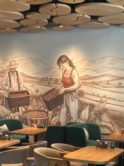 This is an indoor photograph of a cafe with a mural on the wall. The mural depicts a rural landscape with mountains in the background and two figures harvesting what appear to be coffee beans. The figures are a man wearing a hat and suspenders and a woman wearing an apron. Both are holding baskets full of the harvested beans. The ceiling has a decorative wooden design with circular elements. In the foreground, there are several wooden tables and green velvet seating areas, typical of a modern cafe. Small condiment bottles and menus are visible on the tables. The lighting appears to be warm and ambient. This cafe is located in the city of Kokshetau, Kazakhstan.