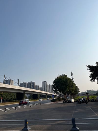The image depicts an urban landscape in Jiangjin, China, under a clear, bright blue sky. In the immediate foreground, a black metal barrier with three grey horizontal bars spans the lower edge. To the right, a dark grey asphalt parking area contains several parked motorcycles and scooters, some equipped with large bundled packages. To the left of the parking area, a multi-lane asphalt road carries traffic, including a red SUV, a white van, and at least two light-colored sedans. A blue "P" parking sign is visible on the roadside.

Across the mid-ground, an elevated concrete structure, serving as a railway or monorail track with overhead catenary wires, traverses horizontally. Supporting pillars are visible beneath the structure. Trees with green foliage are distributed throughout the scene, particularly lining the road and partially obscuring elements. To the right of the elevated track and behind a cluster of trees, a tall, multi-tiered cellular or telecommunications tower stands. In the background, numerous modern high-rise residential buildings with multiple stories are visible behind the elevated track. The far right features an undeveloped, slightly elevated grassy area with sparse vegetation and a visible fence or retaining wall. No individuals are discernibly present. The scene is illuminated by strong sunlight, indicative of morning or late afternoon, casting subtle shadows.