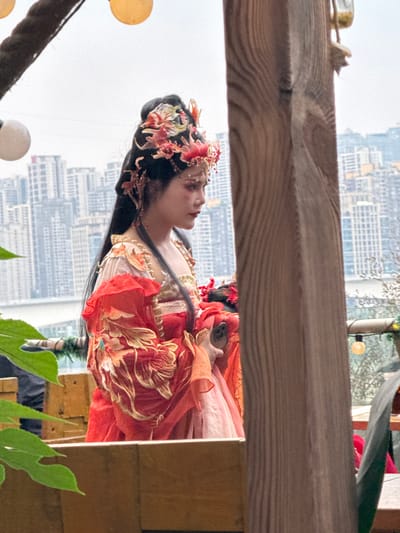 A medium shot depicts a person in traditional Chinese attire against the backdrop of an urban landscape. The person, likely female, has long black hair adorned with floral hair ornaments. She wears a layered, red and cream-colored garment with ornate gold embroidery. The attire suggests a historical or cultural performance context.
The background features tall, densely-packed buildings, typical of a modern city center. The buildings appear in the distance, slightly obscured by haze or atmospheric perspective. The location is consistent with the urban environment of Yuzhong District, China, considering its high-rise architecture and bustling city characteristics.
In the foreground, elements of what appears to be an outdoor set or display are visible, including wooden beams, foliage, and globe-shaped lights. The presence of these elements suggests the subject is posed or situated in a specific environment for a photograph or performance.