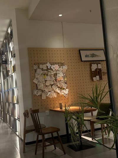 This image depicts an indoor commercial or public space, specifically a quiet corner within the Chongqing High-tech Zone, China. The scene is devoid of people. On the left, a tall, multi-shelved bookcase, illuminated vertically, displays numerous books; a black sign with white text "艺术 ARTS" is affixed to one shelf. The central background features a light-brown wooden pegboard wall. A large black wire grid is mounted on this pegboard, densely covered with multiple small, folded paper notes, indicative of a community message board. To the right, a smaller wooden plank board also holds a few pinned notes. Above these boards, a horizontally oriented framed artwork, likely a landscape painting, is hung. In the mid-ground, a wooden table with a black metal base is positioned, flanked by two wooden chairs; one chair has a light-colored cushion. A white electrical outlet is visible on the wall beneath the pegboard. A green potted plant with long, variegated leaves occupies the immediate foreground on the right, partially obscuring the table and chairs. The ceiling is white and features recessed spotlights. A vertical, translucent fluted glass panel defines the rightmost edge of the frame. The overall environment presents a clean, organized, and inviting atmosphere, potentially a cafe or reading area.