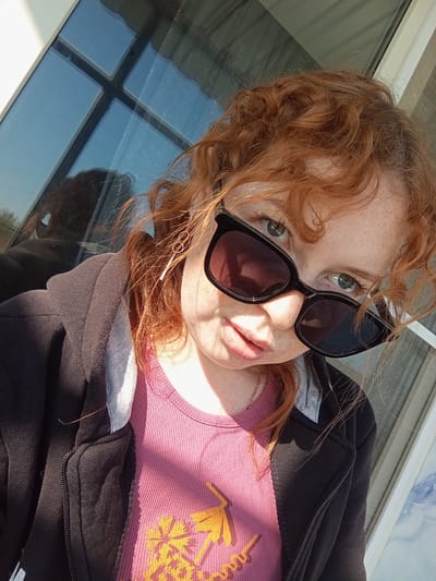 A selfie-style image shows a fair-skinned person with freckles, wearing dark sunglasses, a pink t-shirt with a yellow palm tree graphic, and a partially zipped-up black hooded sweatshirt. The person has red, curly hair and is positioned at an angle in the frame. The background includes a window with visible reflections. The location in Yurgamysh, Russia is unverifiable from the image. No specific actions or interactions are discernible.