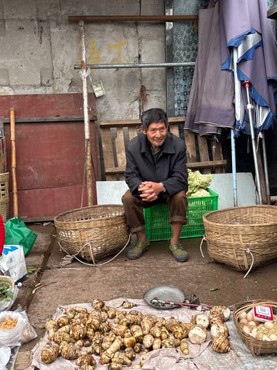 A man is seated cross-legged on the ground in what appears to be a market setting in Jiangjin, China. He is wearing a dark jacket and brown pants with green shoes. He is surrounded by baskets of produce, including eggs and taro root vegetables, some of which are spread out on a cloth. He is sitting in front of a closed door with some crates behind him. A weighing scale is visible near the produce. The environment is a mix of concrete, wood, and metal, suggesting a makeshift market or roadside stall.