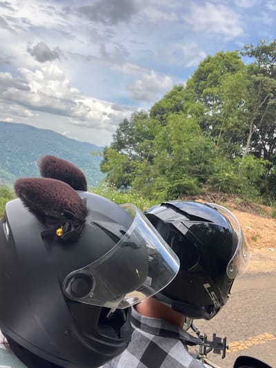 Two people wearing helmets are on a motorcycle, likely in Son Dien, Vietnam, based on the lush green hills and cloudy sky. The helmet in the foreground is black and decorated with brown furry "cat ears" and a small gold bell. The second helmet, to the right, is also black and has a clear visor. The background features rolling green hills covered in vegetation, with a dense cluster of trees to the right. The sky is partly cloudy with a mix of grey and white clouds. The ground appears to be a road with some loose gravel and a yellow line marking. The overall scene suggests a scenic ride through a rural or mountainous area.