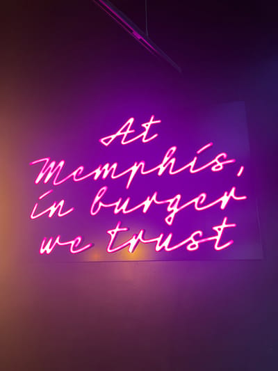 The image displays a vibrant neon sign mounted on a dark wall, glowing with a bright magenta or pink light. The sign features cursive text arranged in four lines. The text reads:
"At this,"
"memphis,"
"in burger"
"we trust"
The letters are stylized in a flowing, handwritten script, with the "m" in "memphis" appearing as a lowercase cursive 'm'. The sign's luminescence casts a strong purple-pink glow onto the surrounding dark surface, creating a moody and modern atmosphere. The background is predominantly dark purple, fading slightly towards black in the upper left and a warmer, possibly orange-brown, hue in the bottom right, suggesting additional ambient lighting or reflections. In the bottom left corner of the image (when oriented as seen), a narrow, reflective surface, possibly part of a metallic object like a laptop or a piece of furniture, is visible, catching some of the sign's light. The overall scene suggests an interior setting, likely a restaurant, bar, or entertainment venue, given the reference to "burger" and the stylistic choice of a neon sign.
