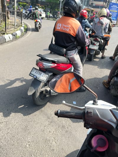 The image depicts a street scene in Bandung, Indonesia. The location is identified as being near the intersection of Jalan Ir. H. Juanda and Jalan Otto Iskandardinata based on the latitude and longitude coordinates. The scene is dominated by a black and white motorcycle in the foreground. The motorcycle is facing the street, and the handlebar is visible. The rearview mirror reflects an orange jacket worn by a person on a red motorcycle. The motorcycle has a license plate that reads "D 2298 ZFH." In the background, there are several other motorcycles and pedestrians. The street is paved and there is a sidewalk to the left of the frame. Several buildings can be seen in the background. The sky is partially obscured by tree branches, indicating that it is daytime and likely a partly cloudy day. The environment appears to be an urban area with busy traffic and multiple vehicles. The image captures a moment in the daily life of Bandung, showcasing the presence of motorcycles and pedestrian movement within the city.