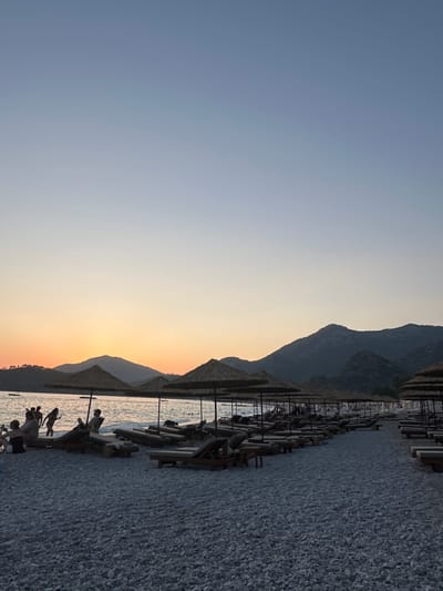 The image depicts a tranquil beach scene at dusk in Fethiye, Turkey. The sky transitions from a pale blue at the top to a warm orange hue near the horizon, where a mountain range is silhouetted against the fading light. In the foreground, a pebbled beach is lined with numerous straw umbrellas and empty lounge chairs, indicating a resort or public beach setting. Several people are visible near the water's edge, with some wading in the shallow sea and others seated on chairs. The overall atmosphere is peaceful and serene, with the soft twilight casting a gentle glow over the landscape.