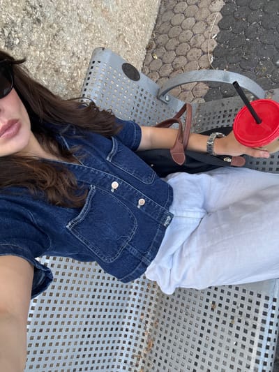 A woman is sitting on a metal bench outdoors, holding a red cup with a straw. She is wearing a denim jacket and white pants. A black bag with brown straps is next to her. The background shows a paved ground with some leaves and dirt. This appears to be an outdoor public space, possibly a park or a street in Tel-Aviv, Israel.