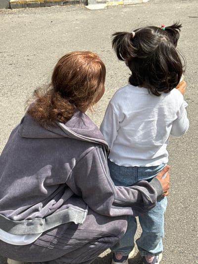 Two individuals stand on an asphalt surface adjacent to a concrete curb in an outdoor environment. The upper individual, a child, has dark hair with two ponytails secured with red and white hair ties, a white ribbed long-sleeved shirt, light blue jeans, and lavender sneakers. The lower individual, possibly an adult, has brown hair and wears a gray zip-up hooded sweatshirt. They appear to be standing still. The location is consistent with an urban environment but specific details to confirm Dmitrov, Russia, are not visible.