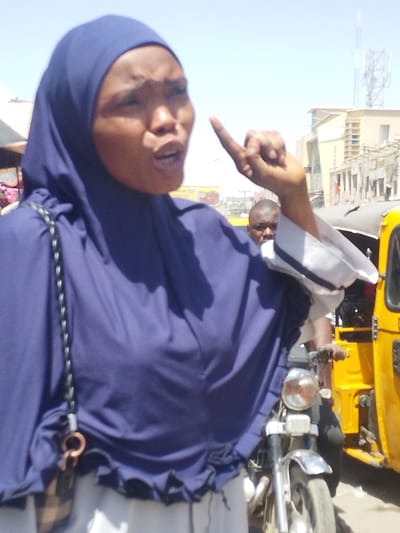 An outdoor scene in Sabon Gari East, Nigeria, depicts a woman in the foreground, positioned slightly left of center, actively speaking or gesturing. She wears a dark blue hijab and a matching top over a white lower garment. Her mouth is open, and her right hand is raised with the index finger pointed upwards. A dark shoulder bag with a plaid pattern and a metal ring is visible on her left side. In the mid-ground, behind the woman, a man with dark skin is partially visible, appearing to be on a vehicle. To the man's right, a yellow auto-rickshaw ("Keke Napep") is partially visible. A motorcycle with a chrome headlight and front fender is situated between the woman and the Keke Napep. The background features several light-colored buildings under a clear, bright sky. A cellular communication tower is visible atop a distant building. Utility poles and lines traverse the background, and what appears to be a commercial sign is partially visible on the left side of the background. The lighting suggests daylight conditions.