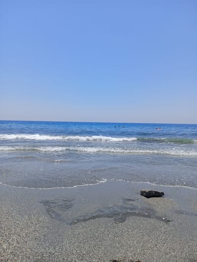 The image depicts a serene beach scene in Alanya, Turkey, under a clear blue sky. The foreground is dominated by wet sand, with gentle waves lapping at the shore. A single, dark-colored rock rests on the wet sand near the center of the frame. In the mid-ground, the clear blue sea stretches towards the horizon, with multiple small, dark figures of people visible in the water, suggesting swimming or other water activities. The horizon line is sharp, separating the calm sea from the expansive sky.