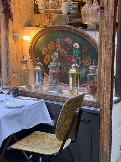 The image depicts a scene inside a restaurant in Paris, France, focusing on details of the interior and table setting.
**Environment:** The interior of the restaurant has a rustic, vintage aesthetic. The walls are textured and painted white or off-white. A wooden beam runs along the ceiling.
**Objects:**
* A large, round, green tray with painted floral designs hangs on the wall, functioning as a decorative element.
* Several glass jars of varying sizes are placed above the tray, containing different food items (nuts, spices, etc.).
* A white table cloth covers the table below, with a simple white plate and silverware on it.
* Several drinking glasses are visible on the table.
* A wooden chair with a black metal frame is positioned near the table.
* A vintage-style light bulb in a white ceramic holder is mounted on the wall.
* Dried red peppers hang near the bottom left corner.
* A crystal pitcher is hung on a wall on the left of the image.
**Subjects:** There are no people visible in the frame.
**Actions:** The scene appears static, depicting a table set for a meal, but currently unoccupied.
**Interactions:** There is no apparent interaction. The objects are arranged in a decorative and functional way.
**Overall:** The image presents a carefully styled scene in a Parisian restaurant, emphasizing vintage details and a refined atmosphere.