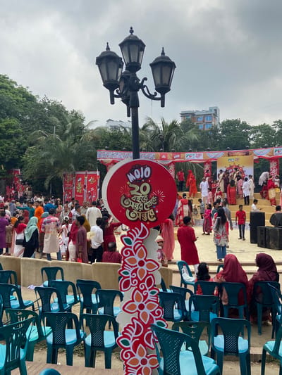 The image depicts a cultural event, likely celebrating Pohela Boishakh, the Bengali New Year, in Dhaka, Bangladesh. A stage is set up with performers in traditional attire, engaging in a musical or dance performance. A crowd is seated in rows of blue plastic chairs, watching the performance. Red and white decorative elements, including a sign with Bengali script, are prominently displayed. A lamppost stands to the side. Palm trees and buildings are visible in the background, suggesting an outdoor location in an urban area. The sky is overcast.