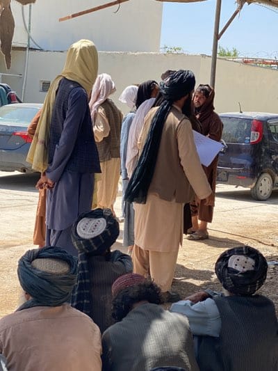 A group of men stand and sit outside in a sunny, outdoor setting, likely a public space in Kandahar, Afghanistan. Most are wearing traditional clothing, including tunics, vests, and turbans or head coverings. Some men stand in a line, while others sit on the ground. One man is holding a document, potentially explaining something to another man. Several vehicles are parked nearby, indicating an urban or semi-urban setting. The presence of turbans and traditional clothing is consistent with the cultural context of Kandahar.