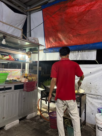 A person, wearing a red shirt and light-colored pants, is seen from behind, cooking at a food stall. They are holding a spatula over a pan placed on a portable stove. The stall itself is a metal cart with a glass display, filled with various food items, bottles, and containers. A red and white checkered cloth hangs on the side of the cart. Behind the stall, there's a backdrop of white and red tarps, suggesting an outdoor or covered market setting. A gas hose is visible connected to the stove. The ground appears to be paved with bricks. The scene is lit by artificial lights, indicating it is likely evening or night. The overall atmosphere seems to be that of a busy street food vendor operation in Denasri Kulon, Indonesia.
