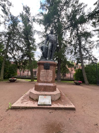 A bronze statue of a man in military uniform stands on a granite pedestal in an outdoor park setting. The statue depicts a man with a mustache, wearing a double-breasted coat with epaulets and trousers, holding a globe in his left arm. The pedestal is hexagonal and made of red granite with inscriptions and a crest. In front of the pedestal, on a lower granite base, is a rectangular plaque. The park features tall trees with green foliage, a dirt path, and a red brick building with a fence in the background. Two flower pots with colorful flowers are placed on either side of the pathway in front of the statue. A lamppost is visible to the right of the statue. The sky is cloudy. This is the monument to Admiral Ivan Fyodorovich Kruzenshtern in Kronstadt, Russia.