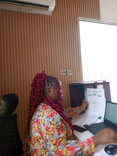 The image depicts a woman with red curly braided hair wearing a floral print top, seated at a desk in what appears to be an office setting. She is holding and examining documents. A laptop computer and a computer mouse are visible on the desk. The documents in her hands have the "Living Spice Nature Cure Hospital" logo visible on them, indicating a medical facility. The office has a vertically-striped wood-paneled wall and a window. An air conditioner is mounted high on the wall. Based on the presence of Living Spice Nature Cure Hospital, the location is Gwarinpa, Nigeria.