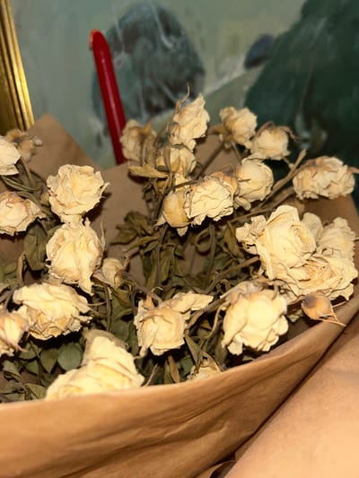 The image shows a close-up of a bouquet of dried, pale yellow roses. The roses are tightly bunched together, with some petals appearing brittle and curled. Their stems and leaves are dry and brown. The bouquet is wrapped in brown paper, which is also somewhat creased and textured. In the background, out of focus, is a painting or tapestry with muted green and dark blue tones, and a thin, bright red candle is positioned vertically to the left of the bouquet. A golden frame is partially visible on the far left edge of the image. The lighting is dim, creating shadows and highlighting the textures of the dried flowers and paper. There are no people or animals visible in the image. The scene does not provide any specific information that would allow for its location to be identified within the city of Plovdiv, Bulgaria.