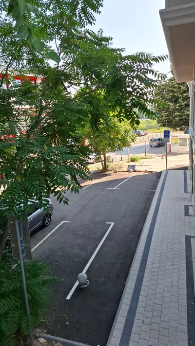 The image depicts a street scene in Plovdiv, Bulgaria, with a focus on a paved parking area adjacent to a building. A portion of a building with a cream-colored facade and white trim is visible on the right side of the frame. A sidewalk made of interlocking gray and black rectangular pavers runs alongside the building.

The parking lot is paved with asphalt and marked with white lines indicating parking spaces. Several of these white lines are clearly visible, forming angled parking bays. In the foreground, a gray sedan is parked in one of the bays. A metal signpost with a rectangular sign is also present near the parking spaces.

Overhanging the parking area from the left is a dense green tree with numerous leaves. In the background, across the street, there are more trees, a body of water, and a few parked cars. A small, booth-like structure with a sign that reads "NON STOP" is situated further down the street, along with a blue and white traffic sign. The sky is clear and blue, suggesting it is daytime.

The scene is devoid of any people. The overall impression is one of a quiet urban environment, possibly residential or commercial, captured from a slightly elevated perspective, looking down into the parking lot and street.