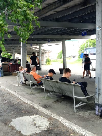 A group of individuals are situated at what appears to be a bus station or transport hub in Pavlovsk, Russia. In the foreground, four people are seated on a long, weathered metal bench under a covered structure. Two young males, one in a black t-shirt and the other in an orange t-shirt with a striped shirt visible underneath, are looking at their phones. An older male with grey hair, wearing an orange t-shirt and patterned shorts, is looking to his left. Another individual, partially obscured, is seated on the far left of the bench. Behind the seated individuals, a man in a black t-shirt walks away from the camera. In the middle ground, a white bus is parked. The environment consists of a paved ground with debris and a concrete curb. The covered structure above is supported by columns, with signs visible indicating platform numbers. Green foliage hangs from branches in the upper left corner of the image.