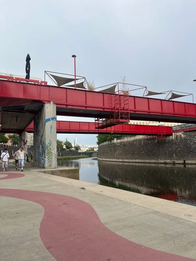 This image depicts a view along the Mže River in Pilsen, Czechia, under an overcast sky. A distinctive red metal bridge structure, with a lower, narrower span beneath a wider upper span, dominates the scene. The bridge is adorned with triangular sunshades and outdoor lighting. In the foreground, a concrete pedestrian path features a red, curved stripe. Three young men, dressed in casual summer clothing, are walking along this path from left to right. One man in a white t-shirt and shorts is closest to the camera, followed by two men in striped shirts and shorts. To the left of the path, a large concrete pillar supporting the bridge is covered in graffiti. The river, reflecting the muted sky and the surrounding architecture, flows along the right side of the image. The riverbank is lined with a dark stone wall. In the background, buildings and other bridges are visible, with a tall, red and white striped industrial chimney standing out against the sky.