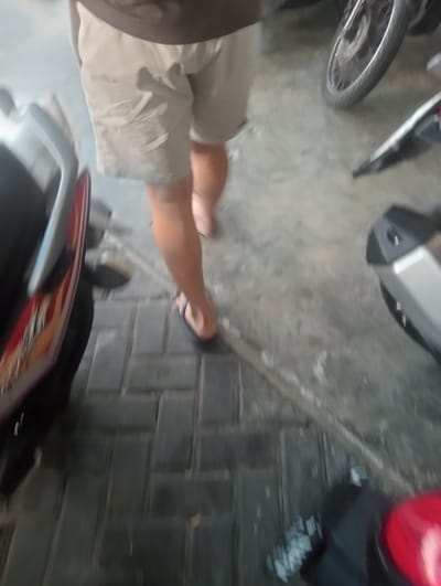 A person wearing tan shorts and flip-flops is walking on a paved surface. The person is walking towards the right side of the image, and their legs and lower torso are visible. Part of a dark-colored motorcycle is on the left, and another motorcycle's wheel is visible in the upper right. The ground is made of interlocking rectangular pavers. The scene appears to be outdoors, possibly in Surabaya, Indonesia, given the style of paving and common motorcycle use.