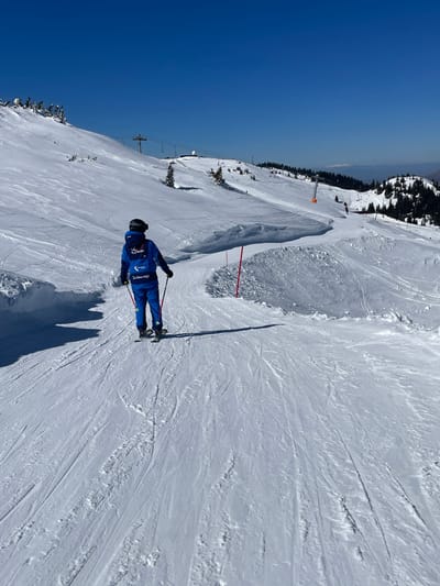 The scene depicts a snow-covered ski slope in Jahorina, Bosnia and Herzegovina, under a clear, cloudless blue sky. A single individual, observed from a rear perspective, is centrally positioned on the groomed slope, equipped with skis and ski poles. The person is attired in a royal blue full ski suit, consisting of a jacket and pants. The back of the jacket features a white graphic incorporating "La Réunion (île)" and "Région" text, with "La Réunion l'île!" printed beneath. A dark helmet covers the head. The individual's shadow is cast prominently to the left on the snow. The foreground and midground consist of packed snow with numerous visible ski tracks. To the immediate right of the skier, two red poles are vertically planted in the snow, marking a boundary alongside substantial snow mounds. The left side of the slope shows undulating snow and a steeper gradient. In the upper left background, a metal ski lift tower is visible ascending the mountain. Further into the background, a white spherical dome structure is present on a distant, snow-capped peak. Coniferous trees are scattered across the midground slopes and form dense forests on the distant mountain ranges. The scene is brightly illuminated by direct sunlight, casting distinct shadows.
