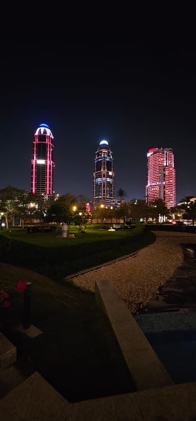 This media file captures a nighttime scene in Doha, Qatar, featuring illuminated skyscrapers and a landscaped park area. Three distinct skyscrapers are prominent, each with unique architectural features and lighting. The topmost building is illuminated with horizontal bands of red light, creating a vibrant, almost fiery effect. Below it, a cylindrical tower glows with blue and white lights, highlighting its glass facade and multiple floors. The bottommost skyscraper mirrors the design of the top one, also adorned with bright red lighting that outlines its structure.
The foreground of the image shows a meticulously maintained park with lush green grass and a winding path covered in light-colored gravel. A concrete structure, possibly a planter or a retaining wall, runs along the edge of the park. In the background, beyond the skyscrapers, faint lights suggest the presence of a city at night. A few trees are visible, their branches silhouetted against the dark sky, and some streetlights cast a warm glow.
The overall mood is serene and sophisticated, indicative of a modern urban environment after dark. The time of day is clearly night, and the clear sky suggests fair weather. The presence of contemporary architecture and landscaping points to a developed city location. No people are visible in the frame, focusing the attention solely on the architectural marvels and the tranquil park. There is no visible text in the media file.