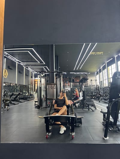The image captures a wide-angle, mirrored reflection of a modern gym in Mengwi, Indonesia. The gym features a sleek, dark color scheme with striking geometric lighting fixtures on the ceiling. In the foreground of the reflection, a woman is seated on a weight machine, holding her phone to take a selfie. She is wearing a black t-shirt and shorts, with white sneakers. To her right, another woman in a red tank top is visible in the background, also engaged with her phone. The gym is equipped with various weight machines and racks of dumbbells, suggesting a well-appointed facility. The overall ambiance is one of a clean, contemporary fitness space.