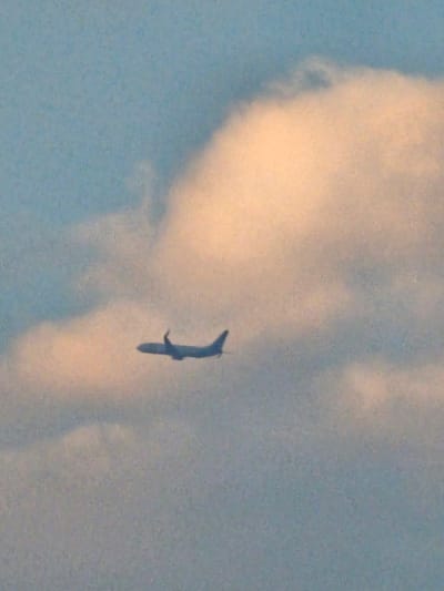 An airplane is silhouetted against a hazy sky with clouds. The airplane is flying from left to right and appears to be in mid-flight. The sky is a light blue with some white and pinkish clouds. The overall image is slightly blurry. There is no discernible location within Denpasar, Indonesia visible.