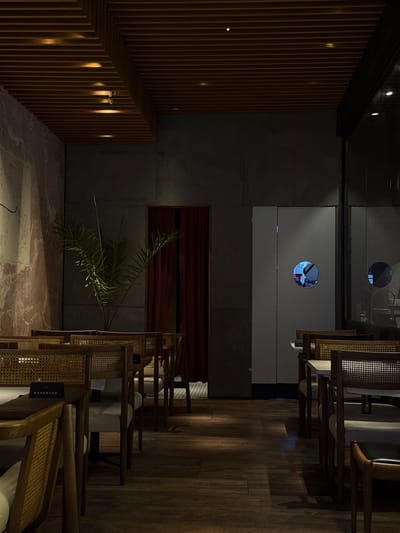 The image depicts the interior of a dimly lit restaurant or cafe located in Yerevan, Armenia. The space features dark wooden plank flooring and a ceiling constructed from parallel wooden slats, embedded with multiple circular spotlights that cast warm light. The left wall has a textured, possibly marbled, reddish-brown and grey finish, while the back wall is a solid dark grey.

Numerous tables with light-colored tops and chairs are arranged in rows throughout the room. The chairs are crafted from dark wood with woven cane backs and seats, some also featuring light-colored upholstered seats. A black "RESERVED" sign rests on a table in the foreground on the left.

In the center-back, a tall, leafy green plant, resembling a palm, is situated in a vase or pot. To the left of the plant, a recessed doorway or archway is covered by a dark red curtain. To the right of the plant, a set of white double doors, likely leading to a service area, features a circular porthole window. Reflected in this porthole is the upper body of an individual, possibly a staff member, wearing light-colored attire and a cap or hat. Further to the right, a large glass partition or window reflects interior elements, including a similar circular shape. No other individuals are directly visible within the dining area, which appears empty.