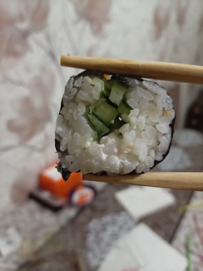A close-up shot shows a single piece of sushi being held by a pair of wooden chopsticks. The sushi roll is cylindrical and appears to be made with white rice, a dark seaweed wrapper, and a filling that includes visible pieces of cucumber and a creamy white substance, likely cheese or a creamy sauce. The background is blurred, but it suggests an indoor setting with a light-colored, textured surface, possibly a tablecloth or bedding. To the left and out of focus, there is an orange object and a hint of purple, possibly part of a food container or condiment. The lighting is soft, and the overall impression is of a casual meal being prepared or eaten. The image has a shallow depth of field, emphasizing the sushi and chopsticks. No text is visible in the image.