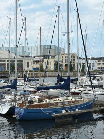 The image captures a harbor scene in Wismar, Germany, with numerous sailboats docked. The foreground features a prominent blue sailboat with a wooden deck and trim, its hull displaying the name "WISMAR". A gangplank extends from the dock to the sailboat. In the background, several other sailboats of various sizes and colors are moored. Behind the boats, industrial buildings with large windows and signage, including "WIS ma e GmbH", are visible. The sky is partly cloudy, and the water reflects the boats and structures. The overall scene suggests a calm day at a German harbor.