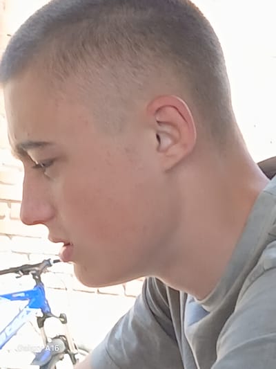 A young male, appearing to be in his late teens, is shown in profile, facing left. He has a closely cropped haircut and is wearing a grey t-shirt. The background consists of a brick wall and a blue bicycle with visible suspension forks. The lower left corner of the image displays the text "Galaxy A16". The image was captured in Ustyvytsia, Ukraine.