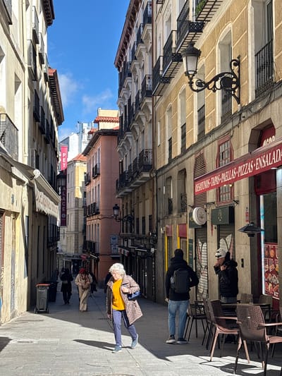 The image displays a narrow, sunlit pedestrian street within a historic district of Madrid, Spain. Tall, multi-story buildings with traditional light-colored facades, wrought-iron balconies, and arched windows flank both sides of the paved street, which recedes into the background under a clear blue sky.

On the right side, a building features an ornate black street lamp and a ground-level establishment with a red awning displaying white text: "DESAYUNOS - TAPAS - PAELLAS - PIZZAS - ENSALADA." An outdoor seating area with dark brown tables and chairs occupies the sidewalk in front of this business. Adjacent to it, another shop exhibits a green cannabis leaf logo sign.

In the mid-ground, an elderly woman with white hair, dressed in a yellow top, patterned coat, and blue pants, walks towards the viewer, casting a long shadow on the pavement. Ahead of her, a man with a black backpack and dark attire walks away. To the right, a person wearing a white beanie and dark jacket stands near the restaurant's seating, holding an object to their mouth. Further down the street on the left, a vertical pink banner with the word "PALACE" is visible. Several other indistinct figures populate the distant background. A black trash bin is positioned on the left side of the street in the foreground.