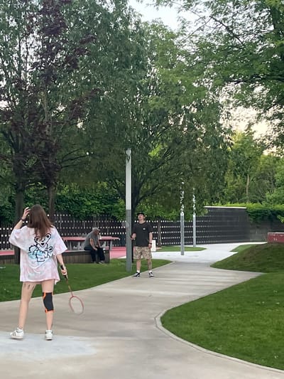The image depicts an outdoor park scene, possibly in Krasnodar, Russia, given the context provided. In the foreground, a young woman with long brown hair is playing badminton. She is wearing a tie-dye t-shirt with a graphic of a bear, shorts, athletic socks, and a black and orange knee brace on her right leg. She holds a badminton racket. Behind her, further down a paved pathway, a man wearing a dark t-shirt and camouflage shorts is standing, also seemingly engaged in playing badminton. In the middle ground, two individuals are seated on a bench near a picnic table. One is wearing a grey t-shirt and a baseball cap, while the other's attire is less clear. The park features lush green grass, mature trees providing shade, and paved walkways winding through the landscape. A dark, slatted fence or wall forms a backdrop behind the seating area. Lampposts are visible along the path, and the overall lighting suggests it is late afternoon or early evening.