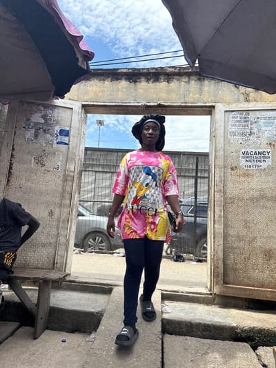 The image shows a woman standing in a doorway, possibly in Lagos, Nigeria. She is wearing a colorful t-shirt with a cartoon character, dark leggings, and black slides. Her hair is covered with a black headscarf. To her left, a person is sitting on a wooden bench, wearing a dark t-shirt and striped shorts. The doorway opens to a street with a metal fence and a car visible in the background. The environment appears to be an outdoor market or a busy urban area, with posters and advertisements on the walls and signs of wear and tear on the structures. The sky is visible above, with scattered clouds.