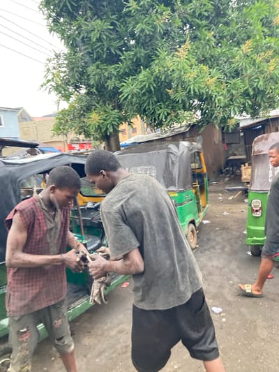 In Umuode, Nigeria, two individuals work on a motorized tricycle (keke). One person, wearing a grey shirt and black shorts, leans over the keke. The other person, wearing a red and black checkered shirt and soiled pants, holds tools. A third individual, wearing a grey t-shirt and green shorts, is visible in the background. The scene takes place in a cluttered roadside environment with buildings and a large tree in the vicinity. Another motorized tricycle is parked behind the one being repaired. The ground is soiled.