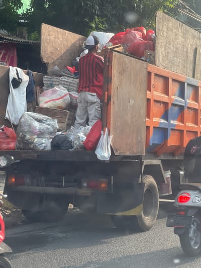 The image depicts a scene in Tangerang, Indonesia, likely a street or roadside. A large, orange and blue truck filled with various types of waste is the central subject. The waste appears to be in plastic bags, some clear and containing visible trash, others opaque red and white bags. There are also some cardboard egg cartons.
A person, seen from the back, is standing in the truck, wearing a red and black striped shirt, light-colored pants, and a white baseball cap. Their posture suggests they might be sorting or arranging the waste.
The truck is on a paved road, with trees and buildings visible in the background, indicating an urban or semi-urban environment. A silver scooter with its brake light illuminated is partially visible in the lower right portion of the frame, suggesting traffic. The overall impression is a utilitarian scene related to waste management in the city.