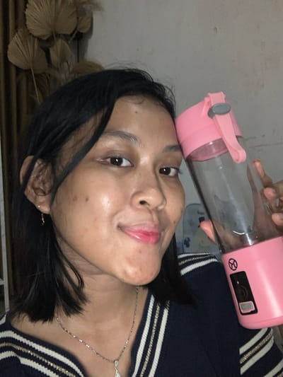 A female subject is depicted in a close-up, indoor setting within Kecamatan Pati, Indonesia, capturing a self-portrait. She possesses dark, shoulder-length hair, styled with a side part and some strands framing her face. Her complexion shows minor blemishes and a mole on her left cheek. She is wearing small gold-colored hoop earrings and a silver-toned necklace with a pendant. Her attire consists of a dark-colored top or sweater featuring horizontal white and gold stripes on the collar/shoulder area. Her expression is a slight smile, with her gaze directed towards the camera. She is holding a clear portable blender with pink top and base sections in her right hand, positioned beside her head. The blender features a grey carrying loop on its lid and a control panel on its base, including a circular symbol with a diagonal line. The background reveals a light-colored wall adorned with several dried, fan-shaped leaves arranged decoratively, and a brown curtain or fabric partially visible on the left. The lighting is subdued.