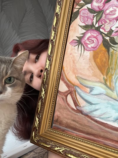 A rotated, low-angle, eye-level shot depicts a painting with a floral arrangement and what appears to be a light-blue bird or angel wing, framed in ornate gold, held aloft to partially obscure a person's face. The person has reddish-brown hair and is looking into the camera. A cat, grey and white, with green eyes, occupies the lower portion of the frame, obscuring the lower part of the person's face. The background is a grey-toned wallpaper with a leafy pattern. The location is not discernible from the image.