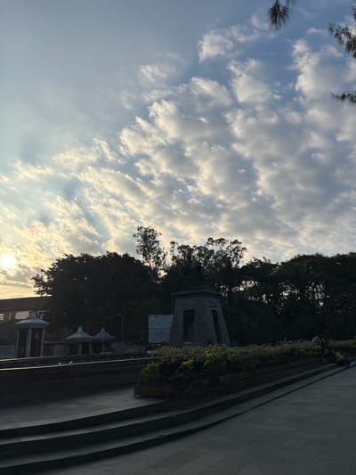 A silhouetted statue stands prominently on a raised pedestal, surrounded by lush, dark green foliage. The sky above is a canvas of white and grey clouds, with streaks of golden sunlight piercing through, suggesting either sunrise or sunset. In the foreground, a series of tiered stone steps leads down from a paved area. To the left, architectural elements with white, pointed roofs are visible, alongside a building with decorative latticework. A few individuals are present in the scene, some casually seated on the steps or walking in the distance, their forms indistinct against the backdrop. The overall impression is one of a serene public space, possibly a memorial or park, bathed in the soft, diffused light of the sky.
