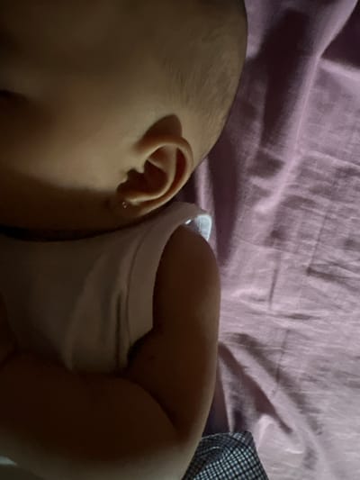 The image shows a close-up, overhead view of a baby's ear and shoulder, lying on a pink textured surface. The baby has fair skin and a small silver earring in its left earlobe. The baby is wearing a white sleeveless top, and a plaid fabric is visible at the bottom edge of the frame. The lighting suggests an indoor setting, likely a bedroom. There is no discernible connection to Lhokseumawe, Indonesia, as the image provides no geographical context.