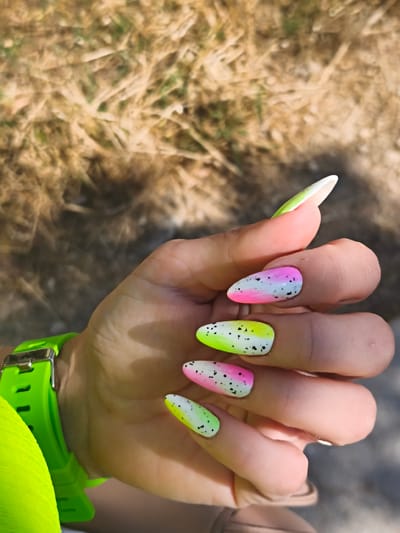 A close-up, eye-level shot shows a woman's right hand adorned with brightly colored, speckled nail art against a blurred background of dry grass and dirt. The nails are long and almond-shaped, each decorated with a gradient of neon green, white, and pink, speckled with black dots. The woman is wearing a neon green watch on her left wrist. The environment appears to be outdoors, possibly a park or a natural area, with the sun casting shadows. The specific location in Sevastopol, Ukraine cannot be determined from the image.
