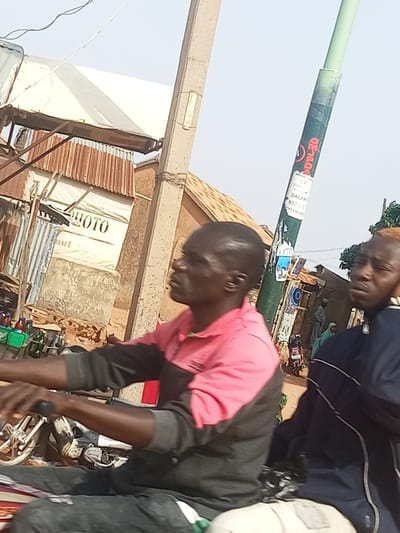 The image depicts an outdoor scene in Arawa, Nigeria, likely a street or roadside market. In the foreground, two individuals are visible, both appearing to be male. The person on the left is in focus, wearing a pink and dark-colored long-sleeved shirt and dark pants. They are seated on a motorcycle, with their hands on the handlebars, suggesting they are either riding or preparing to ride. The person on the right is partially visible, wearing a dark blue jacket with white stripes. They appear to be looking towards the left.
Behind the individuals, there are several structures that suggest a commercial area. A building with a corrugated metal roof and a white awning or sheet is prominent. Text that reads "MOTO" is visible on the white surface, indicating a possible motorcycle repair shop or related business. In front of this building, there are crates containing bottles and other items, consistent with a market stall. A concrete utility pole with various stickers and tape is also a noticeable element in the scene. In the background, other buildings and people in traditional attire are faintly visible, suggesting a populated urban or semi-urban environment. The overall lighting suggests it is daytime, with a bright, possibly overcast sky.