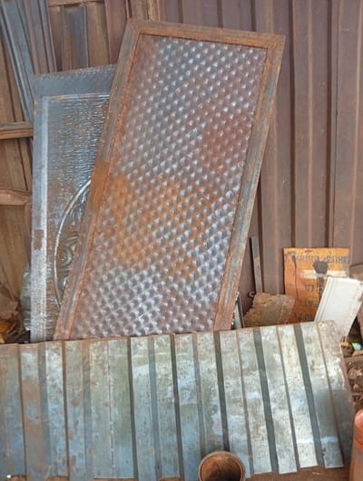 The image depicts a collection of weathered metal materials, suggesting a storage area, scrap yard, or workshop setting in Owerri, Nigeria. The scene is brightly lit, indicating daytime.

In the foreground, a large, horizontal sheet of corrugated metal lies on the ground. It appears worn, with patches of light blue-grey paint or coating mixed with areas of rust and dirt. Below this sheet, a small, rusty, cylindrical metal container or bucket is partially visible.

Behind the corrugated sheet, several other metal panels are propped up. The most prominent is a tall, rectangular panel positioned at an angle, displaying a repeating raised diamond pattern across its surface. This panel is heavily rusted, exhibiting shades of orange, brown, and mottled grey. To its left, a second, wider metal panel, mostly greyish, is partially visible behind the diamond-patterned one. A section of this panel reveals an elaborate, possibly embossed or cast, circular decorative motif. Further left, other narrow metal pieces or beams lean against the background.

The background consists of a vertical corrugated metal structure, likely a wall or fence, also showing signs of rust and weathering. To the right of the central panels, a piece of cardboard or similar material with an orange-brown hue is propped up. Visible text on this material includes a sequence of numbers starting with "0803" and the partial word "EATE". Small, irregular pieces of rusty metal scrap are scattered on the ground around the base of the propped materials. The overall impression is one of disused or awaiting-reuse industrial materials.