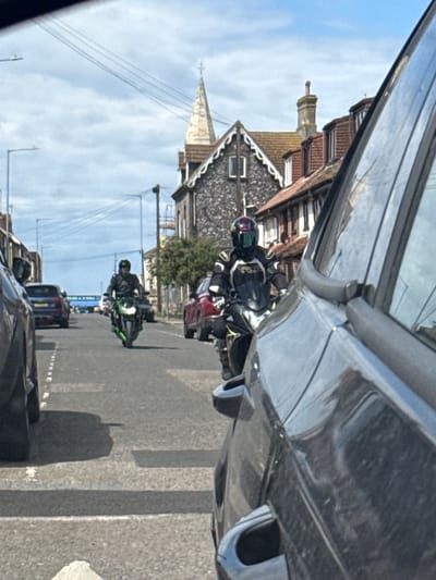 The image, shot from inside a vehicle, captures a street scene in Great Yarmouth, United Kingdom. A convoy of motorcyclists rides along a road lined with buildings on either side. The motorcycles are varied in color, with green and red examples visible. The road is marked with painted lane dividers. A variety of other vehicles are also visible on the road. Buildings of brick, stone, and tile feature in the background, exhibiting details such as pointed roofs, chimneys, and decorative gables. Overhead, utility lines traverse the sky, which is lightly clouded. The interior of the car in which the image was taken is a prominent element of the foreground, partially obscuring the view.