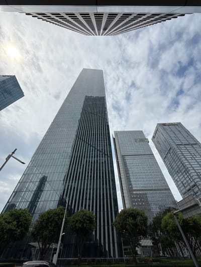 This is a wide-angle, low-angle photograph of a cluster of modern skyscrapers in Tianhe District, China, on a cloudy day. The sun is visible through the clouds on the left side of the frame, casting a slight glare. The buildings are tall and appear to be made of glass and steel, with a variety of architectural styles, including a prominent building with a reflective, angled glass facade and another with a grid-like pattern of windows. The foreground shows the tops of several green trees, suggesting a street-level view looking upwards. A lamppost with a modern design is visible on the left. The overall impression is one of a bustling, contemporary urban environment.