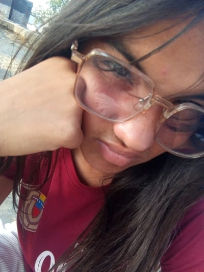 A close-up selfie depicts a woman with long, dark hair, resting her chin on her hand. She wears eyeglasses with a transparent frame. Her shirt is maroon and features a patch that includes the colors of the Venezuelan flag. The background is somewhat blurry, revealing some hints of an outdoor environment, possibly a street or paved area with some greenery. The location cannot be determined to be Juan Griego, Venezuela from the available visual information.
