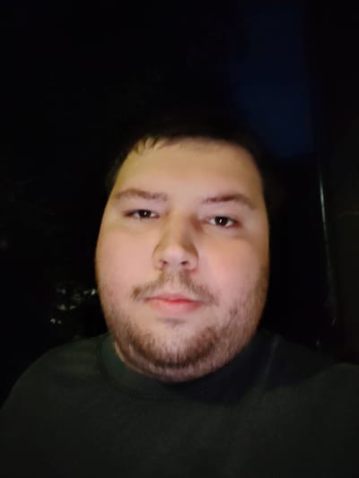 The image is a close-up, head-and-shoulders selfie of a man taken at night. He has light brown hair, brown eyes, and a light beard stubble. He is wearing a dark green crew neck shirt. The background is dark, suggesting an indoor or nighttime outdoor setting. There is no indication of the city of Vladimir, Russia, or any specific location within it.